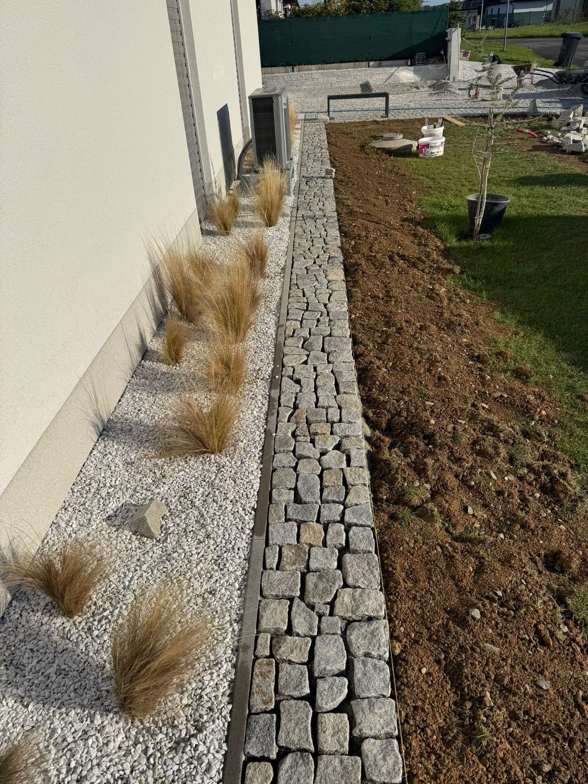 Okrasný záhon z bílého štěrku s travinami lemovaný žulovou kostkou podél domu