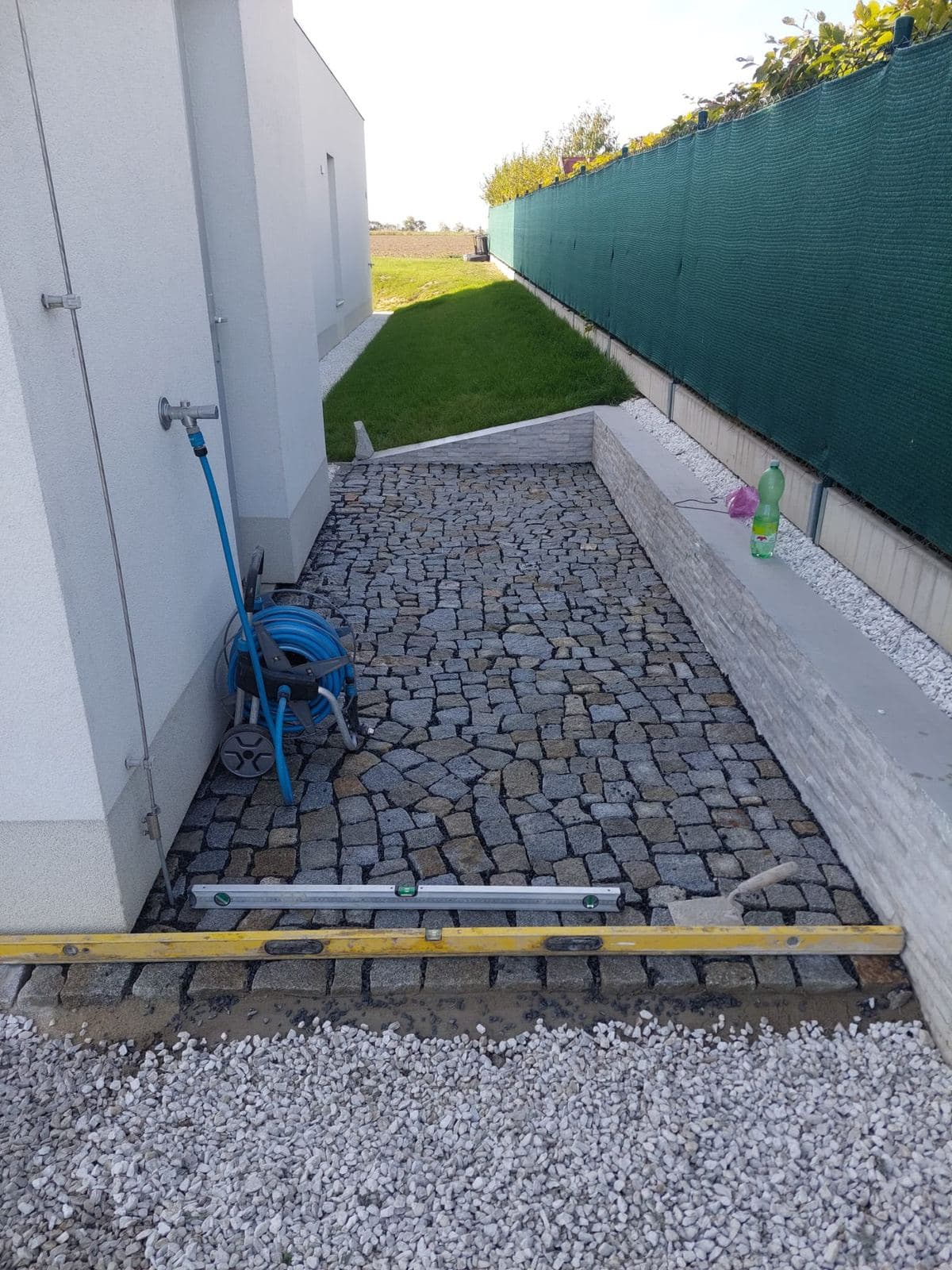 Pokládka chodníku ze žulové kostky podél bílé fasády s opěrnou zídkou